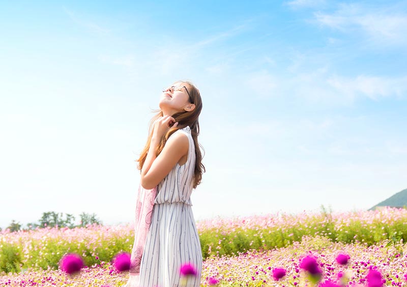 Woman in a field of pink flowers looks toward the blue sky, eyes closed, and inhales deeply.